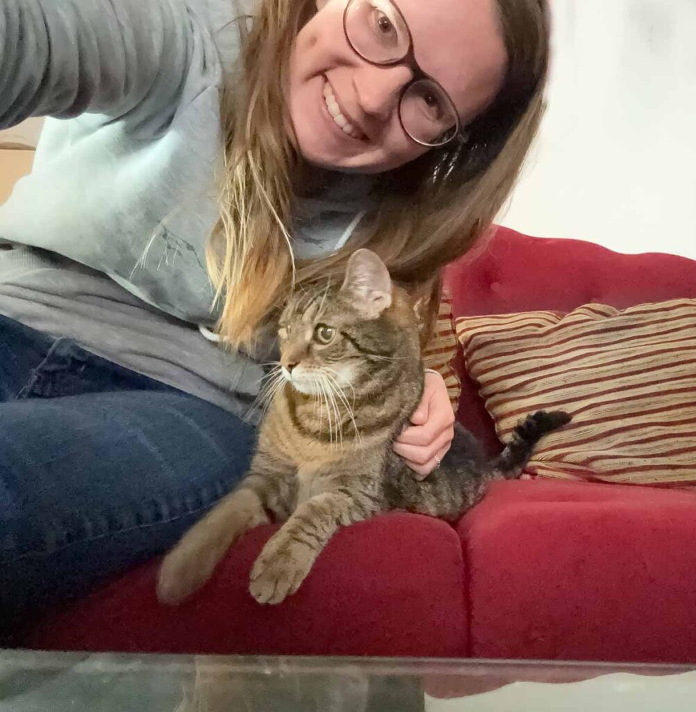 Anna Hopfengärtner sitzt mit ihrer getigerten Katze auf dem roten Sofa und lächelt in die Kamera – die SEO-Expertin in gemütlicher Homeoffice-Atmosphäre.